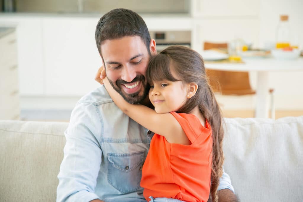 A heartwarming moment as a father is embraced by his daughter on the sofa, showcasing the bond between them.