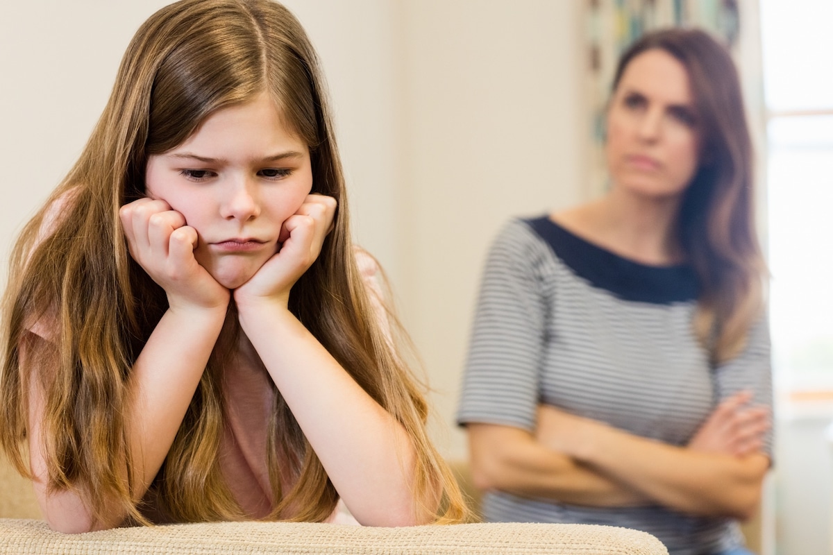 Upset daughter sitting in the living room, with her mom in the background.