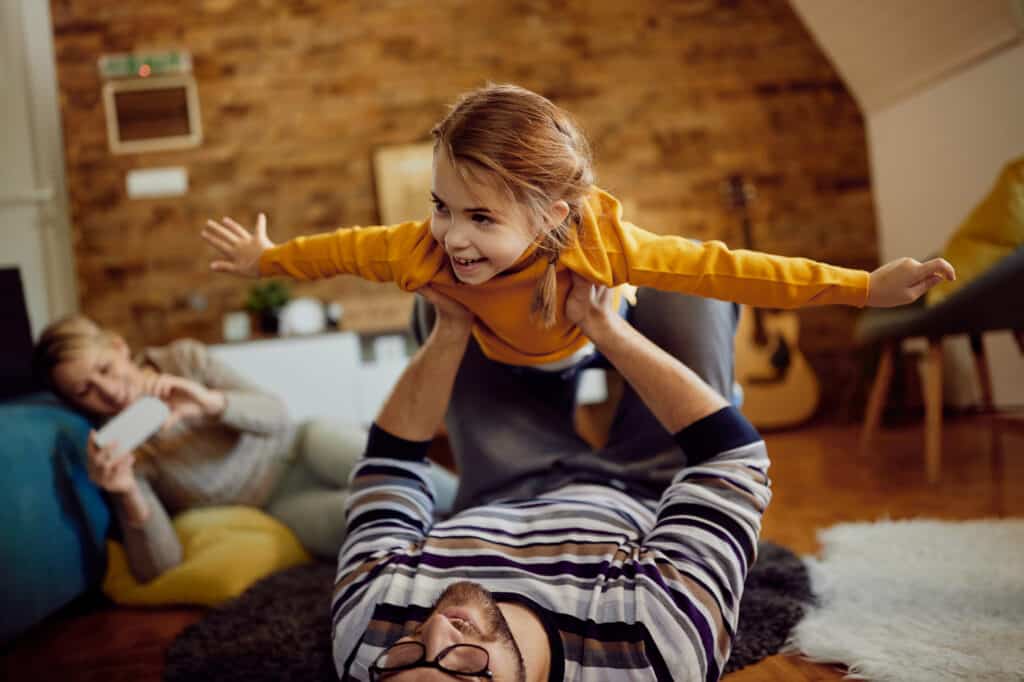 A joyful moment captured as a father playfully holds his daughter imitating an airplane, while the mother captures the moment with a photo.