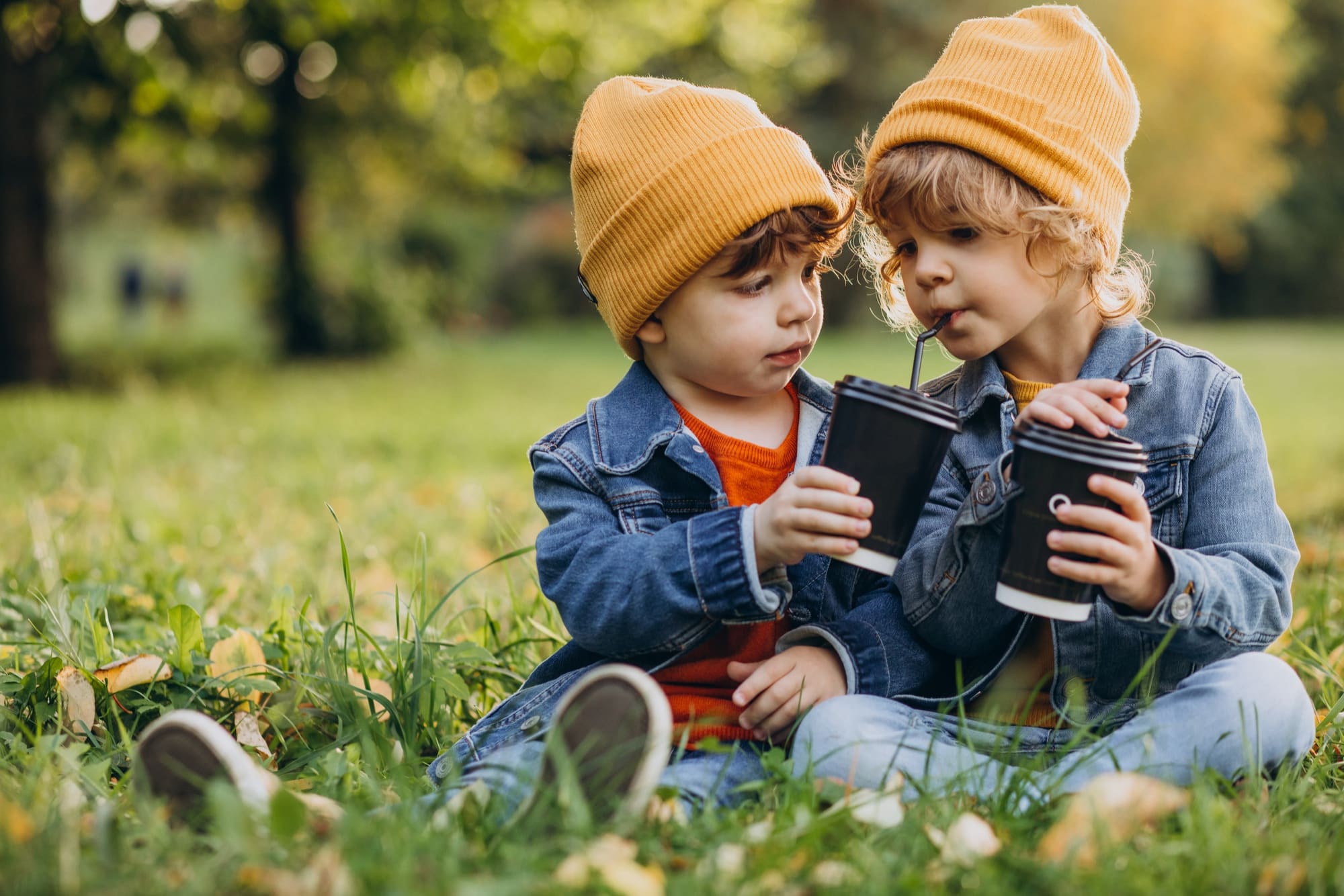 Two little brothers sitting on the grass, happily sharing cups of hot chocolate.