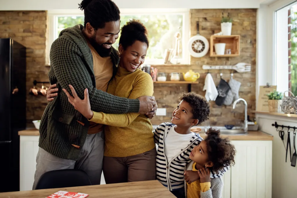 Mom and dad share a warm embrace in the kitchen as their two little kids watch with joyful smiles