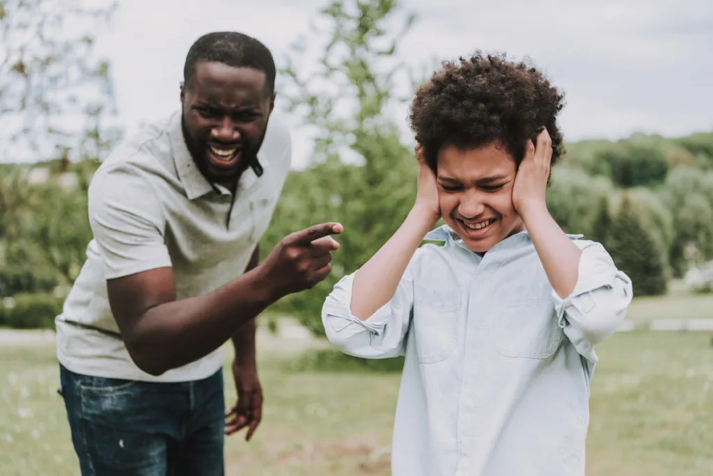 child rearing - Angry dad yelling and pointing a finger to his scared school-aged son that is covering his ears 