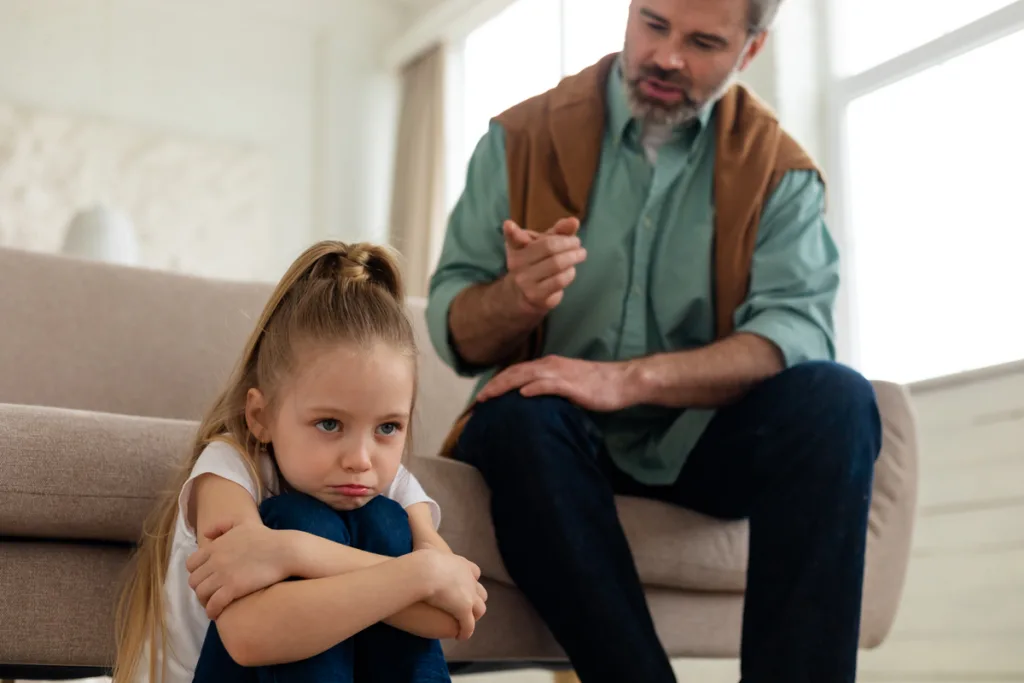 Dad sitting on the sofa scolding young daughter who is sitting on the floor hugging her knees