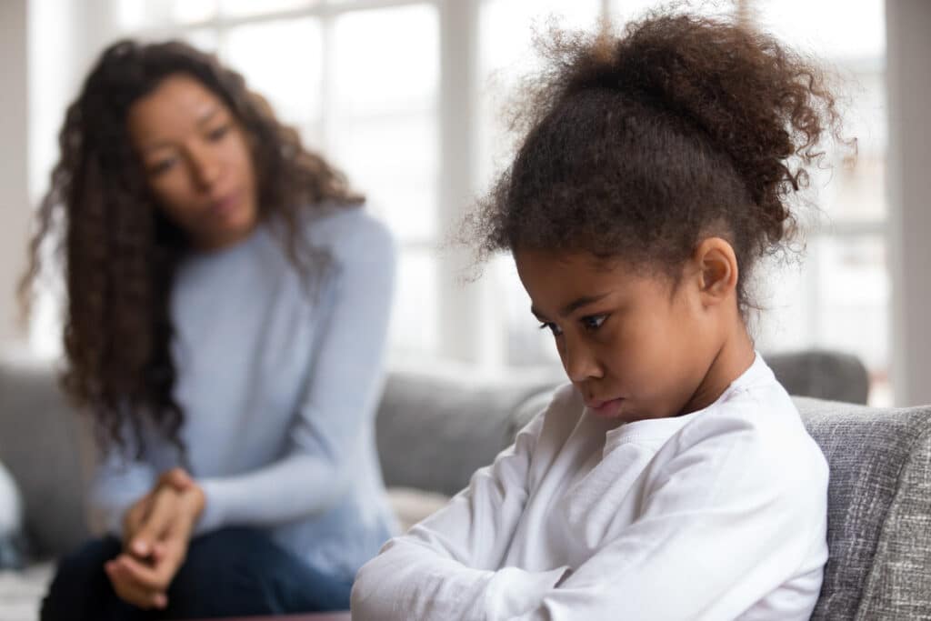 A displeased little girl with crossed arms next to her mother on the couch