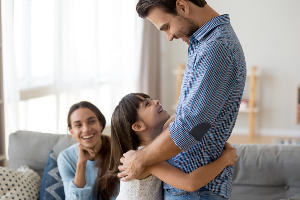 A little girl lovingly wrapping her arms around her dad’s waist while smiling and making eye contact