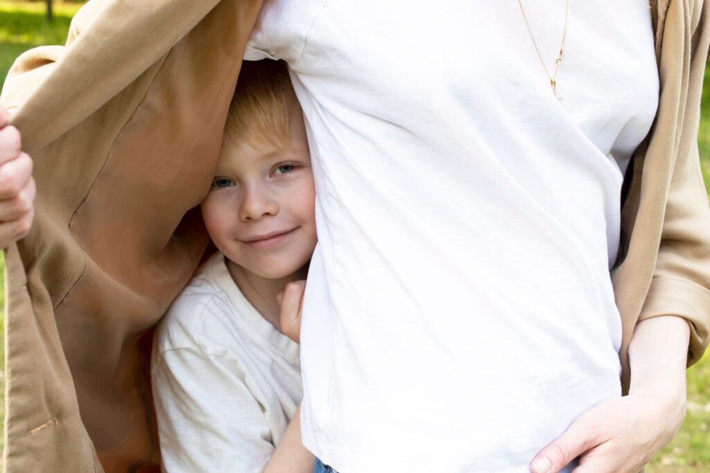 A smiling child hiding under their mother’s coat