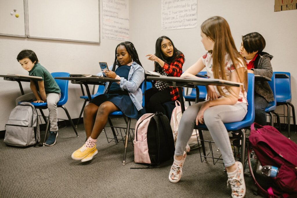 Young girl holding phone in classroom, being pointed at and laughed at by other kids.