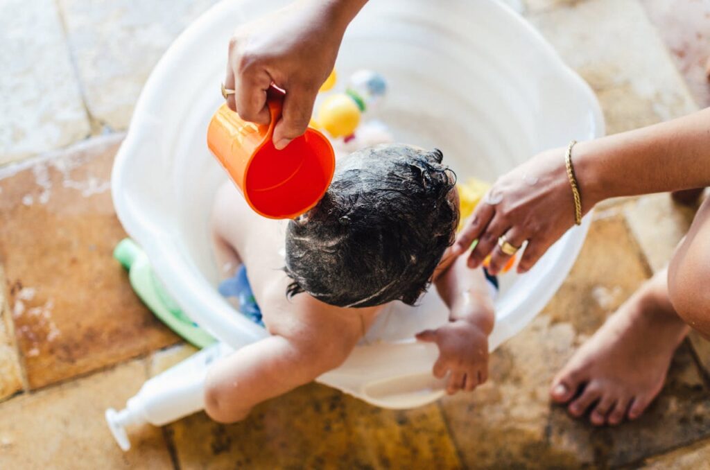 A toddler bathing in a basin outdoors, with his mom rinsing his hair using a mug.