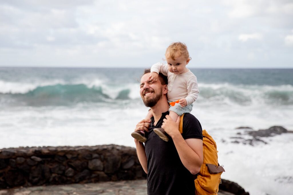 Father walking on the beach with toddler on his shoulders, as the child playfully hits him.